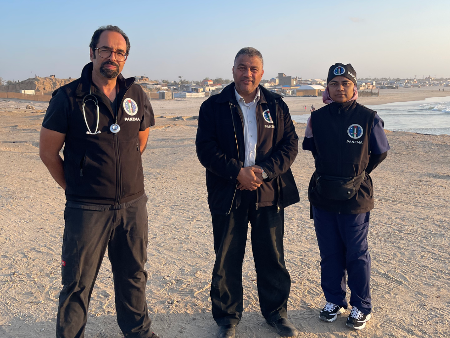 Three people standing on a beach, dressed in clothes with PANZMA patches, and have stethoscopes and scrubs.